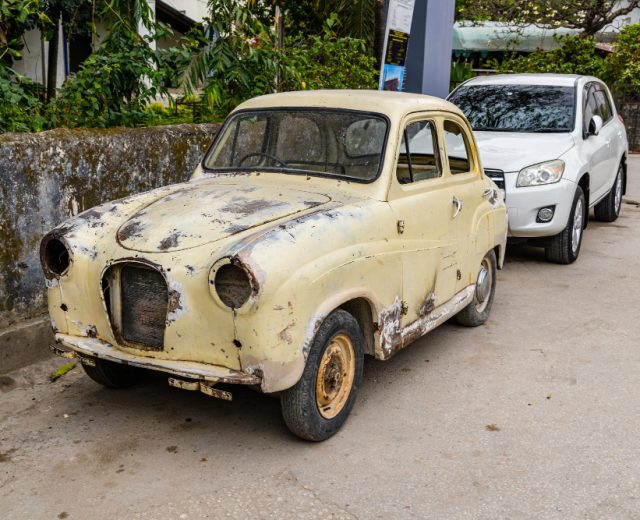 An old, rusted car is parked in front of a white SUV on a street, with greenery and a stone wall in the background, reminding one of the necessity for vehicle disposal services.