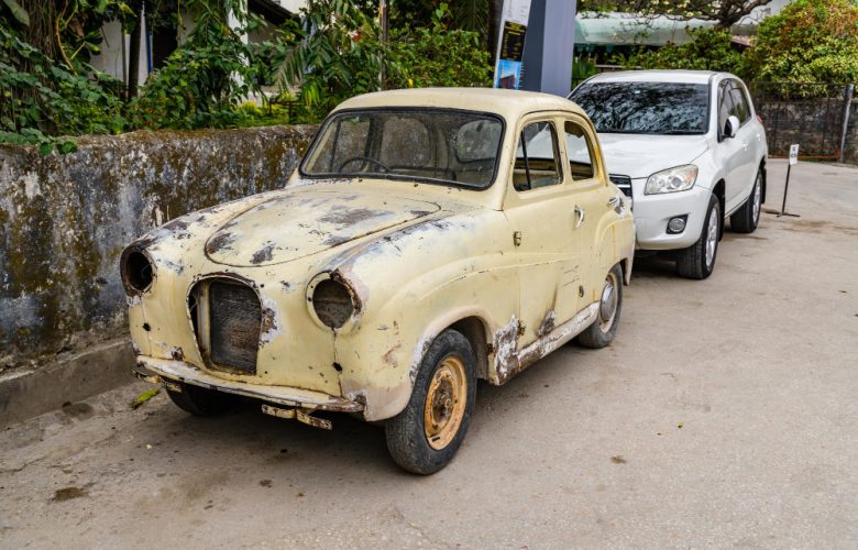 An old, rusted car is parked in front of a white SUV on a street, with greenery and a stone wall in the background, reminding one of the necessity for vehicle disposal services.